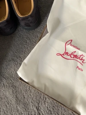 Svarta sneakers från Christian Louboutin - Snygga svarta sneakers från Christian Louboutin i mocka med coola svarta nitar på tån. Skorna har rund tå, platt sula och snörning. Insidan är beige och de har en lyxig vibe som sticker ut. Perfekta för dig som vill ha något unikt och edgy.