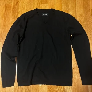 Zadig & Voltaire V-neck - Svart v-ringad stickad tröja från Zadig & Voltaire med långa ärmar och mörkgrå förstärkningar på armbågarna. Tröjan har diskreta ribbade muddar och en klassisk passform som funkar till många olika stilar. Perfekt för dig som gillar stilrena och enkla plagg. 100% merino