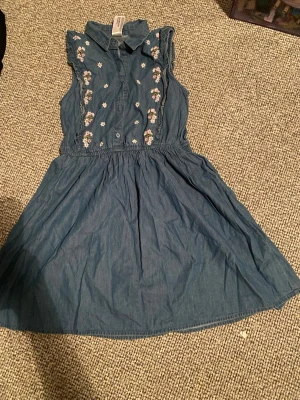 Blå ärmlös klänning från Palomino - Supersöt blå kortklänning från Palomino med krage och knappar framtill. Klänningen är ärmlös med volangdetaljer vid axlarna och har broderade blommor i rosa och vitt längs bröstet. Tillverkad i mjuk bomull, perfekt för varma dagar.Fina mönster 