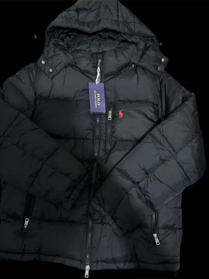 Svart pufferjacka från Polo Ralph Lauren - Säljer en svart pufferjacka från Polo Ralph Lauren med huva och dragkedjor. Jackan har det klassiska röda Polo-logot på bröstet och flera praktiska fickor med dragkedja. Perfekt för kalla dagar och riktigt snygg till streetwear-stilen.