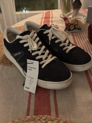 Moncler sneakers - Snygga sneakers från Moncler i mörkblå mocka med vita detaljer och snörning. Skorna har rund tå och platt sula, samt vit logotyp på hälen. Insidan är fodrad i vitt skinn och yttersulan är vit med Moncler-märkning. Perfekt för dig som gillar stilrena och exklusiva sneakers. Självklart äkta och kollade med experter hos vinted, verifierade!