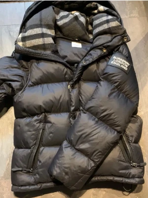 Burberry jacka - Svart pufferjacka från Burberry med klassisk logotyp på bröstet och rutigt foder i huvan. Jackan har dragkedjor på fickorna och en rejäl huva. Perfekt för kalla dagar och har en modern, oversized look.