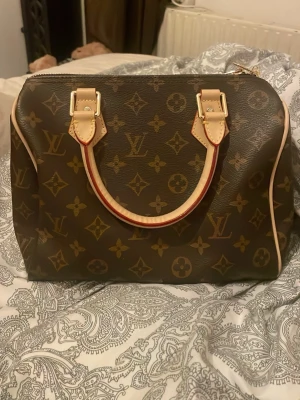 Monogram handväska - Säljer en klassisk handväska i brunt monogrammönster med beigea handtag och detaljer. Väskan har guldfärgade metalldetaljer och dragkedja. Aldrig använd. Möts upp i Stockholm eller söder om Stockholm 💕