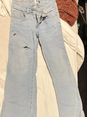 Ljusblå jeans från 15 Paris Code - Säljer ett par ljusblå jeans från 15 Paris Code med slitna detaljer och hål på framsidan. Byxorna har klassisk femficksdesign, dubbla knappar i midjan och dragkedja. Materialet är jeans och passformen är rak med normal midja.