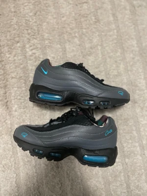 Nike Air Max 95 grå och svarta sneakers - Nike Air Max 95 sneakers i grått och svart med blå detaljer. Ovandelen har mesh och skinn, med vågiga paneler och synlig Air-enhet i sulan. Svart snörning och blå logga på sidan och tungan ger en clean look. Perfekta för dig som gillar streetwear och sportig stil. Skicka gärna frågor och bud!