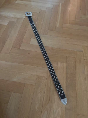 Svart glittrigt bälte med fjärilar - Säljer ett svart bälte med glittrig yta och silverfärgade detaljer. Spännet är dekorerat med strass och har två snygga fjärilar. Bältet har även små runda hål längs hela bandet för extra edge.