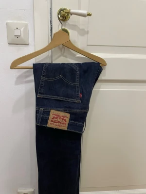 Levi's 505 mörkblå jeans - Säljer ett par klassiska Levi's 505 jeans i mörkblå tvätt. De har raka ben, normal passform och är tillverkade i slitstarkt bomullsmaterial. Jeansens ikoniska läderpatch och röda Levi's-tag syns tydligt på bakfickan. Perfekt för dig som gillar tidlös stil.