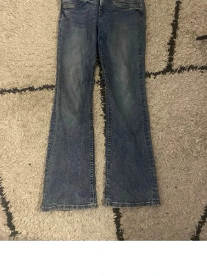 Blå bootcut jeans med slitningar - Snygga blå jeans med bootcut-snitt och klassisk femficksdesign. Jeansen har en tvättad look med lätt slitna detaljer och raka sömmar. Materialet är robust denim som ger en cool vintagekänsla. Perfekt för dig som gillar retrostil och vill ha ett par jeans med lite extra attityd. 