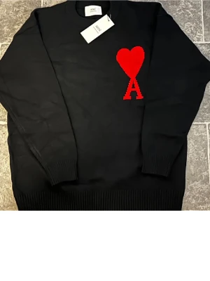 Svart stickad tröja från Ami - Svart stickad tröja från Ami Alexandre Mattiussi med ett stort rött hjärta och bokstaven A broderat på bröstet. Tröjan har rund hals, ribbade muddar och lång ärm. Perfekt för dig som gillar stilrena detaljer med en twist.