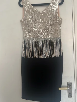 Svart och silver paljettklänning med fransar - Snygg svart klänning med överdel i silverfärgade paljetter och långa fransar som hänger ner från midjan. Klänningen är ärmlös och har en v-ringad hals. Perfekt för dig som vill sticka ut med glittriga detaljer och cool design.