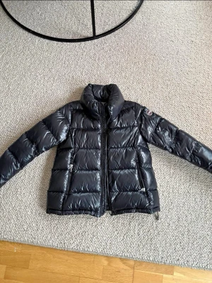 Svart pufferjacka från Colmar - Säljer en svart pufferjacka från Colmar i dun med glansig finish och hög krage. Jackan har två sidofickor med dragkedja och en tydlig logga på ärmen. Perfekt för kalla dagar och har en snygg, modern look. Passar dig som gillar stilrena och funktionella jackor dam.   Knappt använd.  Storlek 42, känns som small/medium.  Personen på bilden med jackan på är 165cm normalbyggd en small/medium.