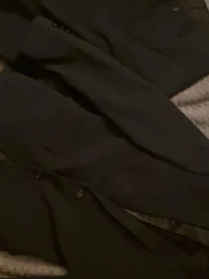 Mörkblå kostym - Snygga svarta kostymbyxor med rak siluett och klassisk design. Byxorna har knapp och dragkedja framtill samt fickor på sidorna. Perfekta för dig som gillar en stilren look och vill ha ett par byxor som funkar till skjorta eller kavaj. Materialet är troligtvis ull eller polyester. Både kavaj och byxor ingår. Dm mig för mer bilder eller frågor 