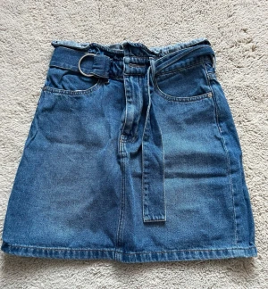 Blå jeanskjol med bälte från H&M - Säljer en snygg blå jeanskjol från H&M i storlek 36. Kjolen har ett medföljande bälte med metallring, två framfickor och två bakfickor. Använd endast en gång. 