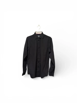 Svart skjorta från John Henric - Svart skjorta från John Henric med klassisk krage och knappar framtill. Skjortan har lång ärm och är tillverkad i mjuk bomull som känns skön mot huden. Perfekt för dig som gillar stilrena och enkla plagg med snygg passform.