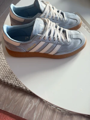 Adidas Handboll Spezial  - Säljer ett par Adidas Spezial sneakers i ljusblå mocka med vita detaljer och klassiska tre ränder på sidan. Skorna har vit snörning och en brun gummisula som ger en retro vibe. Perfekt för dig som gillar stilrena och sportiga sneakers. Dessa skor har inga täcken på användning 