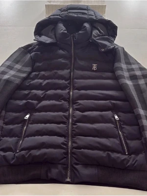 Svart pufferjacka från Burberry S - Snygg svart pufferjacka från Burberry med huva och klassiskt rutigt mönster på ärmar och rygg. Jackan har två fickor med dragkedja, Burberry-logga på bröstet och quiltad framsida. Perfekt för dig som vill ha en stilren och varm jacka med lyxig känsla.