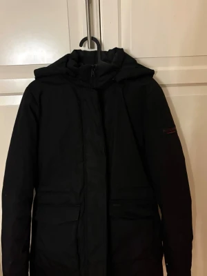 Svart parkajacka från Navahoo - Svart parkajacka från Navahoo med huva och dragkedja framtill. Jackan har två stora fickor med lock och en stilren design som passar perfekt när det är kallt ute. Materialet är polyester och jackan är långärmad med normal passform. Köp nu jag postar snabbt!!💕💕