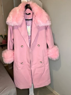 Rosa trenchcoat ,kappa med fuskpäls och pärlknappar och bälte  - Säljer en ljusrosa kappa med bred krage och muddar i fluffig fuskpäls. Dubbelknäppt med stora pärlknappar och två fickor framtill. Insidan är quiltad för extra värme. Perfekt för dig som vill sticka ut med en girly vibe.