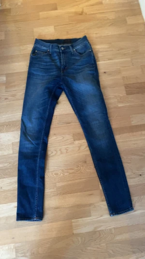 Blå jeans från Tiger of Sweden - Säljer ett par klassiska blå jeans från Tiger of Sweden. Jeansen har fem fickor, normal passform och raka ben. Snygga kontrastsömmar och läderpatch bak med T/J-logga. Perfekta till en chill vardagslook.