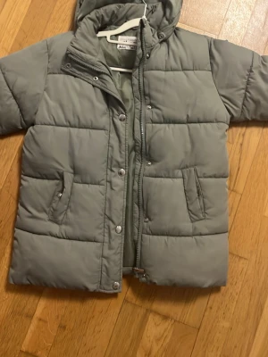 Grön pufferjacka med huva - Säljer en grå pufferjacka från Aid-Health med huva och quiltad design. Jackan har dragkedja och tryckknappar framtill, samt två fickor med knappstängning. Perfekt för kalla dagar och har en clean look som funkar till det mesta.