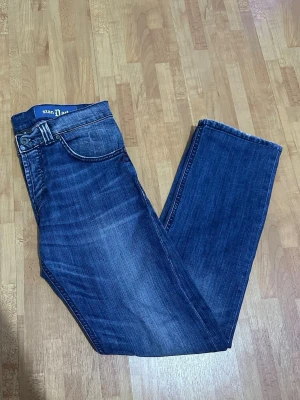 Dondup STYLE UP008 050U - Säljer ett par snygga blå jeans från Dondup med klassisk femficksdesign och raka ben. Jeansen har en mörk tvätt med lätt slitning och detaljerade sömmar på bakfickorna. Perfekt passform och coola detaljer vid linningen.