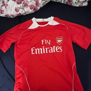 Arsenal Adebayor 25 matchtröja röd - Säljer en röd Arsenal matchtröja med vitt tryck, Fly Emirates-logga och klubbmärke på framsidan. På ryggen står Adebayor och nummer 25 i stora vita siffror. Tröjan har vita detaljer vid kragen och längs sidorna. Perfekt för dig som älskar fotboll och Arsenal.