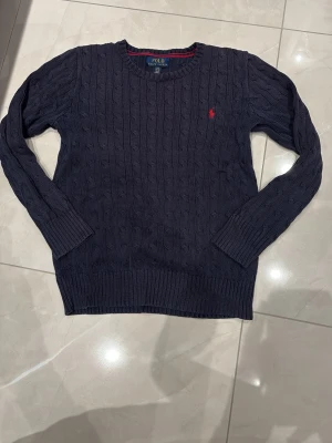 Mörkblå stickad tröja Polo Ralph Lauren - Snygg mörkblå stickad tröja från Polo Ralph Lauren med klassiskt kabelmönster och röd broderad logga på bröstet. Rund halsringning och ribbade muddar vid ärmslut och nederkant. Perfekt för dig som gillar stilrena och ” stockholms stil”  plagg. Denna tröja är i gott skick även fast den är väl andvänd