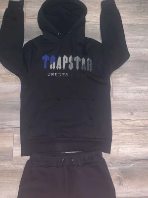 Trapstar dress  - Svart dress från Trapstar med logga i blått och grått och lite svart. Byxorna har snörning i midjan och är tillverkade i mjukt bomullsmaterial som är jätte skön. Passformen är slim och benen är raka med dragkedja vid benslut. Luvan har också en snörre man kan spänna med och det är jätte bra kvalitet och jätte skön. Perfekt för chill eller streetwear. OBS!!!! (PRISET KAN DISKUTERAS UNDER SNABB AFFÄR)