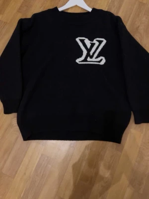 Svart stickad tröja från Louis Vuitton - Svart stickad tröja från Louis Vuitton med stor vit LV-logga på bröstet. Tröjan har rund halsringning och långa ärmar. Perfekt för dig som vill ha en stilren och lyxig look med streetkänsla.