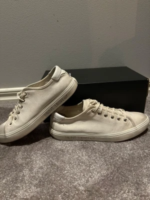 Vita Saint Laurent sneakers i canvas - Säljer ett par stilrena vita sneakers från Saint Laurent i canvas med klassisk låg profil och snörning. Skorna har rund tå, platt sula och Saint Laurent-logga tryckt på sidan av sulan. Metallöljetter och vita detaljer ger en clean look som passar till det mesta.