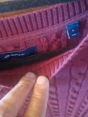 Vinröd kabelstickad tröja från GANT - Säljer en vinröd kabelstickad tröja från GANT i storlek L. Tröjan har rund hals, ribbade muddar och ett broderat GANT-logo på bröstet. Materialet är mjukt och stickat, perfekt för kyliga dagar. Passformen är normal och ärmarna är långa.
