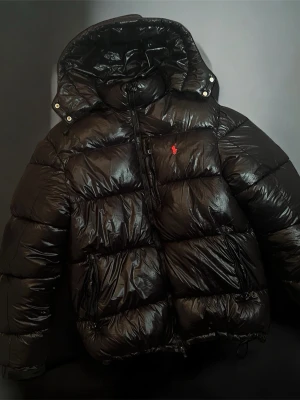 Svart pufferjacka från Polo Ralph Lauren - Svart, glansig pufferjacka från Polo Ralph Lauren med röd broderad logga på bröstet. Jackan har huva, dragkedja framtill och två sidofickor med dragkedja. Perfekt för kalla dagar och har en riktigt schysst streetstil.