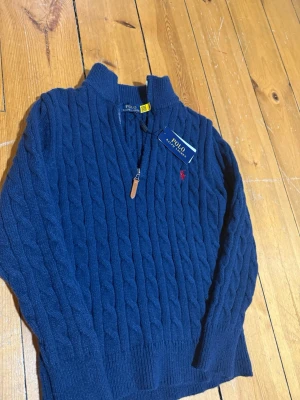 Marinblå stickad tröja Polo Ralph Lauren   - Säljer en marinblå stickad tröja från Polo Ralph Lauren med kabelstickat mönster och halv dragkedja vid halsen. Tröjan har lång ärm, ribbade muddar och en liten röd logga broderad på bröstet. Perfekt för dig som gillar klassisk och stilren design.
