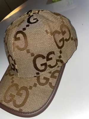 Gucci jumbo  - Säljer en beige keps från Gucci med det klassiska GG-mönstret i brunt. Kepsen har en böjd skärm med brunt kantband och justerbar rem i brunt läder baktill. Materialet är slitstark canvas och detaljerna ger en lyxig känsla.