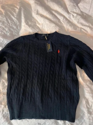 Mörkblå stickad tröja Polo Ralph Lauren - Snygg mörkblå stickad tröja från Polo Ralph Lauren i storlek M men den passar även S. Tröjan har kabelstickat mönster, rund hals och en röd broderad logga på bröstet. Perfekt för dig som gillar klassisk stil med en modern touch.