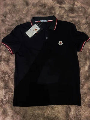 Svart Moncler pikétröja med röd/vit detalj - Svart pikétröja från Moncler med klassisk krage och knappar framtill. Röd och vit rand på krage och ärmslut ger en snygg kontrast. Broderad Moncler-logga på bröstet. Perfekt för dig som vill ha en stilren och sportig look.