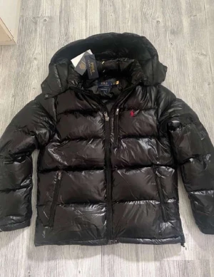 Svart pufferjacka från Polo Ralph Lauren - Säljer en svart pufferjacka från Polo Ralph Lauren med blank finish och röd broderad logga på bröstet. Jackan har huva, dragkedja framtill och två sidofickor med dragkedja. Perfekt för kalla dagar och riktigt snygg streetstil.