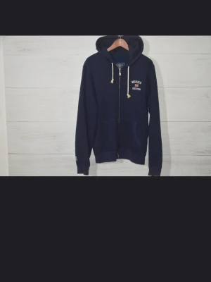 Marinblå zip-hoodie från Morris - Säljer en marinblå hoodie från Morris med dragkedja och huva. Tröjan har vita snören, fickor framtill och tryckt logga på bröstet. Perfekt för chill dagar och enkel att matcha med jeans eller joggers.