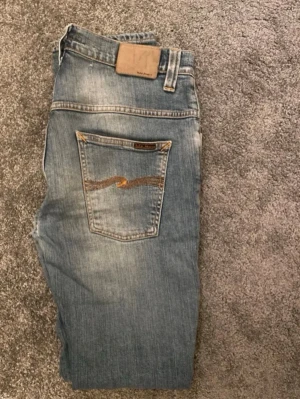 Blå jeans från Nudie Jeans - Säljer ett par  blå jeans från Nudie Jeans Med RIKTIGT bra passform  och klassisk femficksdesign samt snygga slitningar. Jeansen har en broderad detalj på bakfickan och är tillverkade i mjukt jeanstyg med dragkedja och knapp framtill. Kostar 1800+ nypris Pris går att diskuteras 