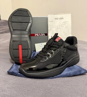 Prada skor - Säljer ett par svarta Prada sneakers med blank finish och meshdetaljer. Skorna har snörning, rund tå och platt sula. Diskret röd logga på plösen och sulan. Materialet är en mix av skinn och syntet, vilket ger en lyxig och modern vibe. Kommer med originalkartong och dustbag.