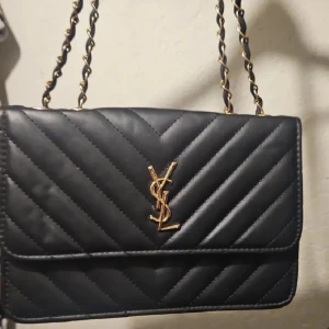 Svart quiltad axelväska från YSL - Snygg svart axelväska från YSL med klassisk quiltad design och guldfärgad kedja. Väskan har YSL-logga i guld på framsidan och är tillverkad i skinn. Perfekt för dig som vill ha en stilren och lyxig accessoar.