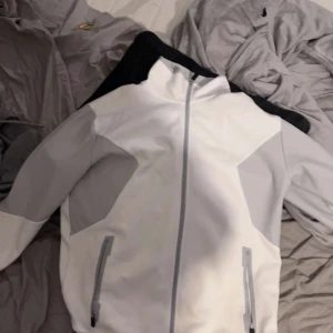 BOSS Vindjacka - Snygg vit och ljusgrå vindjacka från BOSS med hel dragkedja framtill och två fickor med dragkedja. Jackan har en sportig look och är tillverkad i polyester, perfekt för dig som gillar stilrena och moderna plagg.