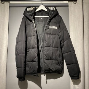 Svart pufferjacka från Hoodrich M - Svart pufferjacka från Hoodrich med broderad logga på bröstet och insidan av huvan. Jackan har dragkedja framtill, huva och quiltad design. Perfekt för kalla dagar och ger en clean streetwear-look.