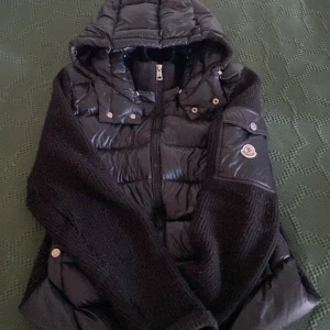 Svart Moncler pufferjacka med stickade ärmar - Snygg svart pufferjacka från Moncler med stickade ärmar och huva. Jackan har dragkedja framtill, quiltade detaljer och en ficka med Moncler-logga på ärmen. Perfekt för kalla dagar och ger en cool streetstyle-vibe.