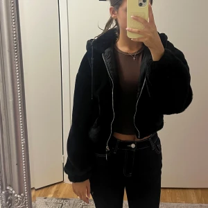 Svart croppad pälsjacka med huva - Säljer en svart, croppad pälsjacka med huva och dragkedja framtill. Jackan har långa ärmar, snörning vid huvan och en oversized vibe. 