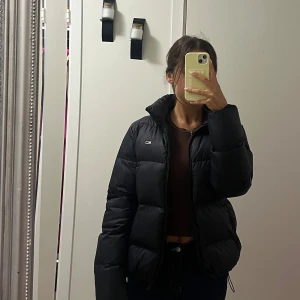 Svart pufferjacka från Tommy Hilfiger - Säljer en svart pufferjacka från Tommy Hilfiger med hög krage och dragkedja framtill. Jackan har en clean look med liten logga på bröstet och breda, vadderade paneler. 