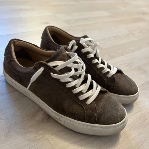Bruna mockasneakers från Les Deux - Snygga bruna sneakers från Les Deux i mocka med vita snören och vit sula. Klassisk låg modell med rund tå och stilren design. Perfekta för dig som gillar en clean och avslappnad look.