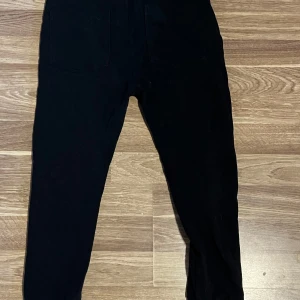 Svarta mjukisbyxor från 157, stl 140 - Svarta mjukisbyxor från 157 med dragsko i midjan och dragkedjefickor framtill. Byxorna har ribbade muddar vid bensluten och är tillverkade i en mjuk bomulls- och polyestermix. Perfekta för chill eller träning.