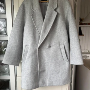 Ljusgrå oversized kappa - Snygg ljusgrå kappa med oversized passform och klassisk krage. Jackan har en enkel knäppning framtill och två fickor på sidorna. Perfekt för dig som gillar stilren och minimalistisk look.