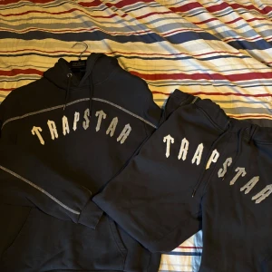 Svart Trapstar dress med tryck - Svart hoodie från Trapstar med stort vitt logotyp-tryck över bröstet och kontrastsömmar. Klassisk huva med snörning och känguruficka framtill. Tillverkad i bomull och polyester för en skön och streetig vibe. Byxor följer med!